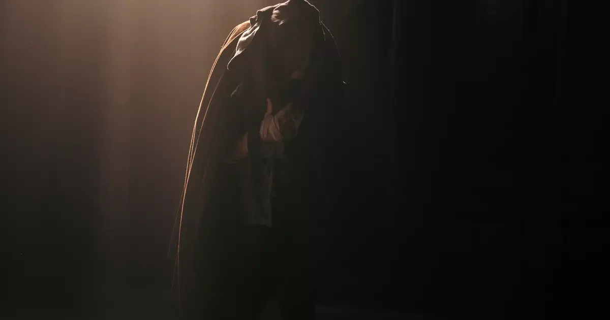 Silhueta encurvada de um homem coberto por uma capa, iluminada por um feixe de luz em cena do filme