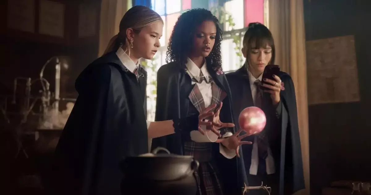 Três estudantes de capa preta observam uma esfera rosa flutuando sobre um caldeirão em cena de Coven Academy