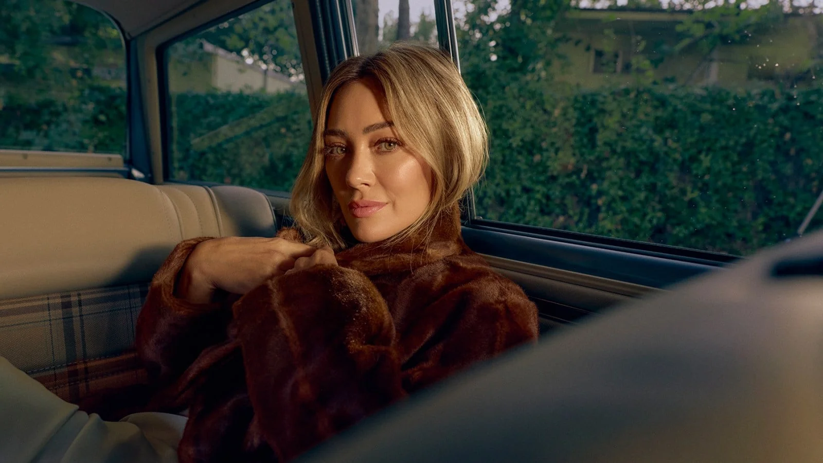 Hilary Duff no banco de trás de um carro, com casaco marrom, em foto de divulgação