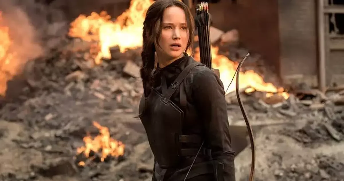 Katniss Everdeen, com arco e flecha nas costas, diante de ruínas em chamas